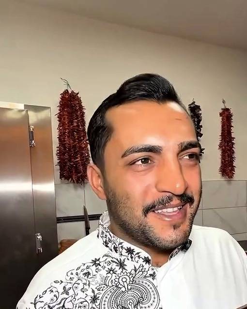 Reşit Usta mutfakta, ocak başında gülümsüyor; arkada duvarda kuru biber dizileri